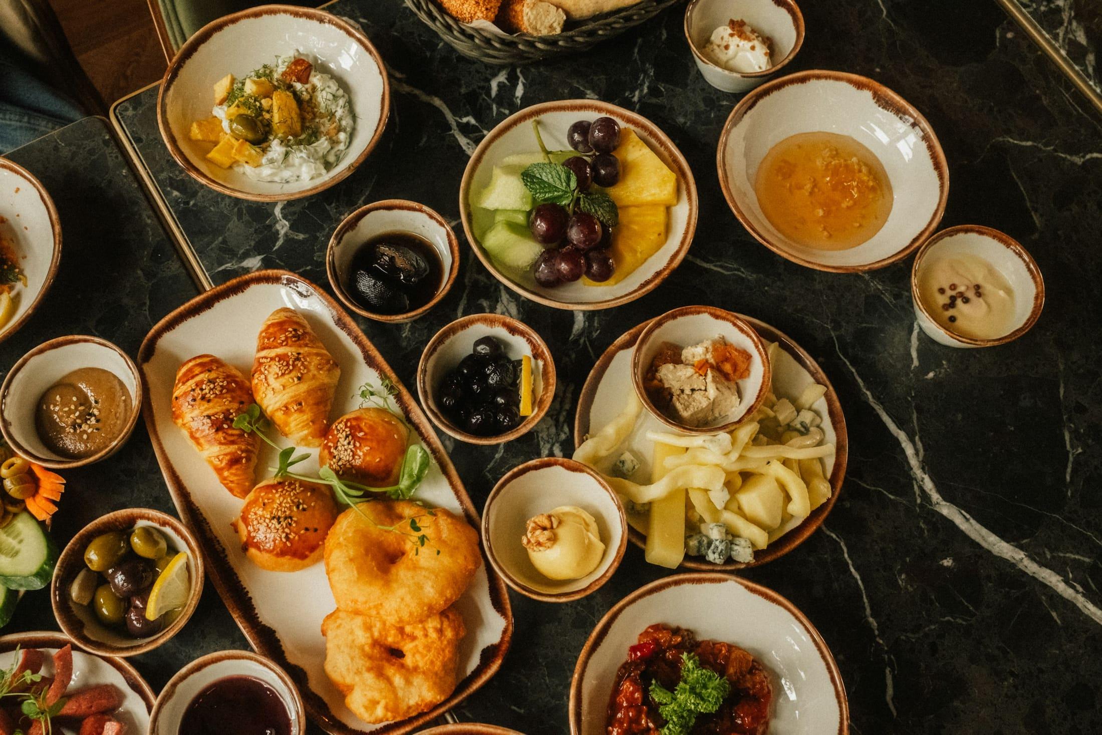 A breakfast table on dark marble, laid with croissants, fresh fruit, cheeses, olives and honey